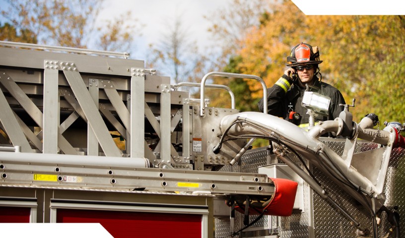 Fireman standing on the back of a firetruck, smiling with phone device in hand