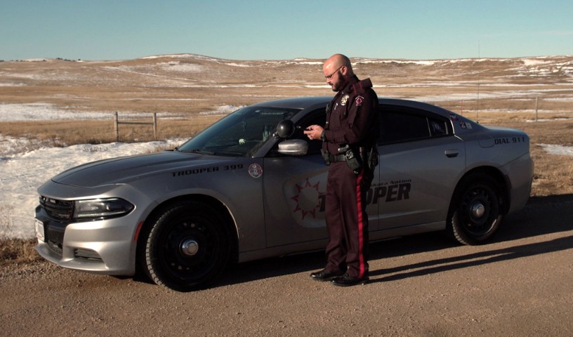 State trooper in snow-covered prairie standing outside of his car on his device