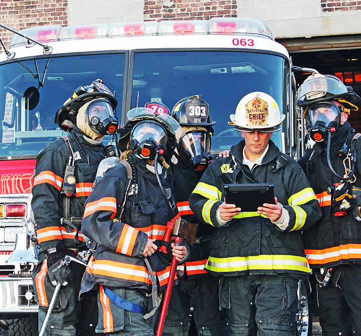 4 firefighters with masks on prepare to engage fire, standing outside of their truck with the Chief outlining their plan of attack on a FirstNet mobile tablet device.