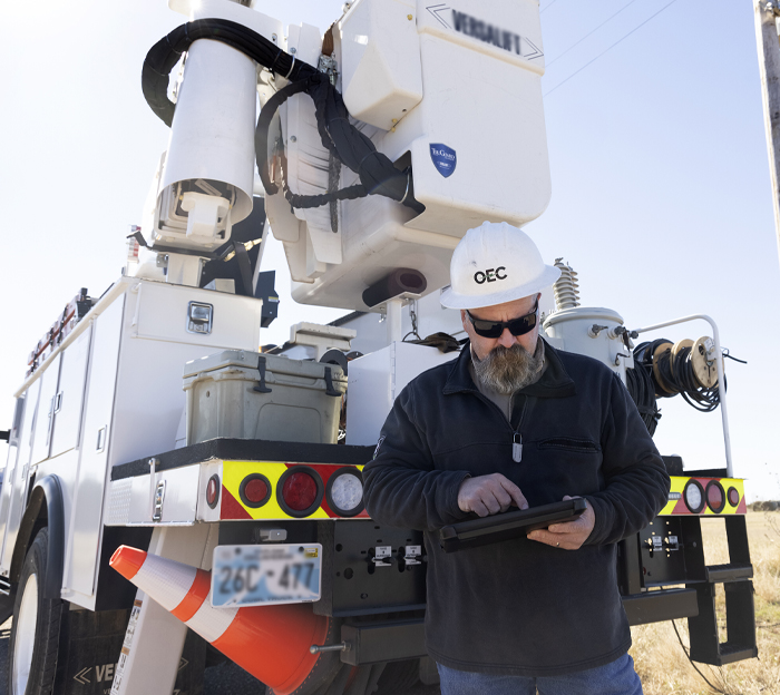 First responder in white hard hat and sunglasses, browsing tablet device behind a lift truck.
