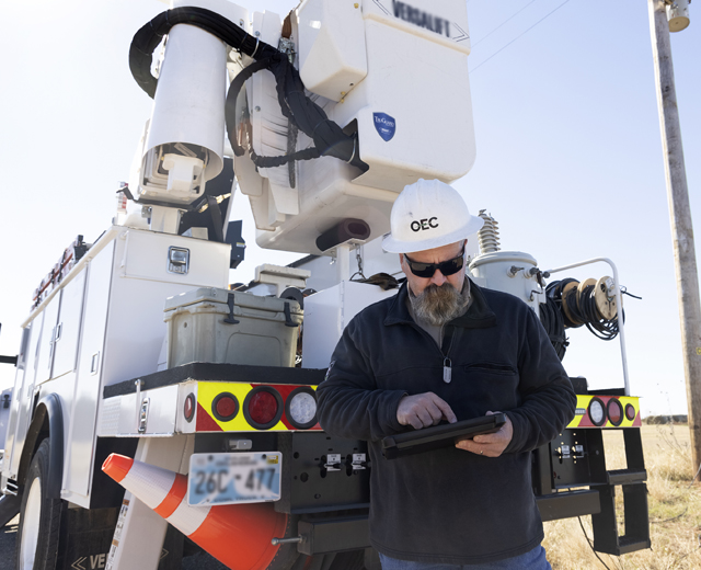First responder in white hard hat and sunglasses, browsing tablet device behind a lift truck.