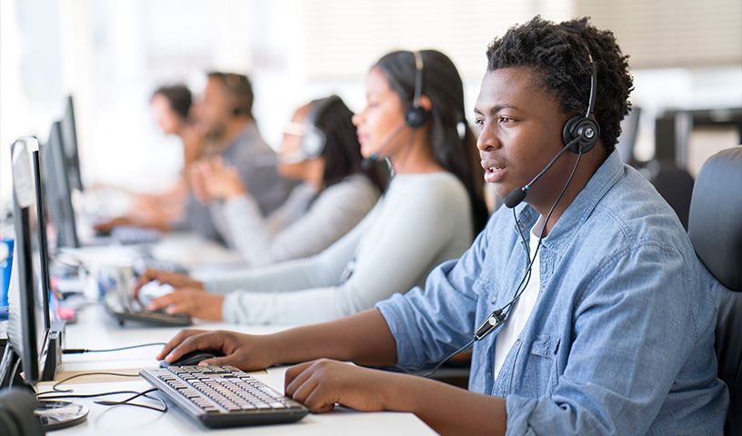 A group of call center employees seated in a row at desks, wearing headsets and working on desktop computers.